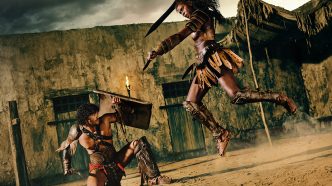 Nick E. Tarabay as Ashur stands in a blood-spattered arena surrounded by roaring gladiators in Spartacus: House of Ashur.